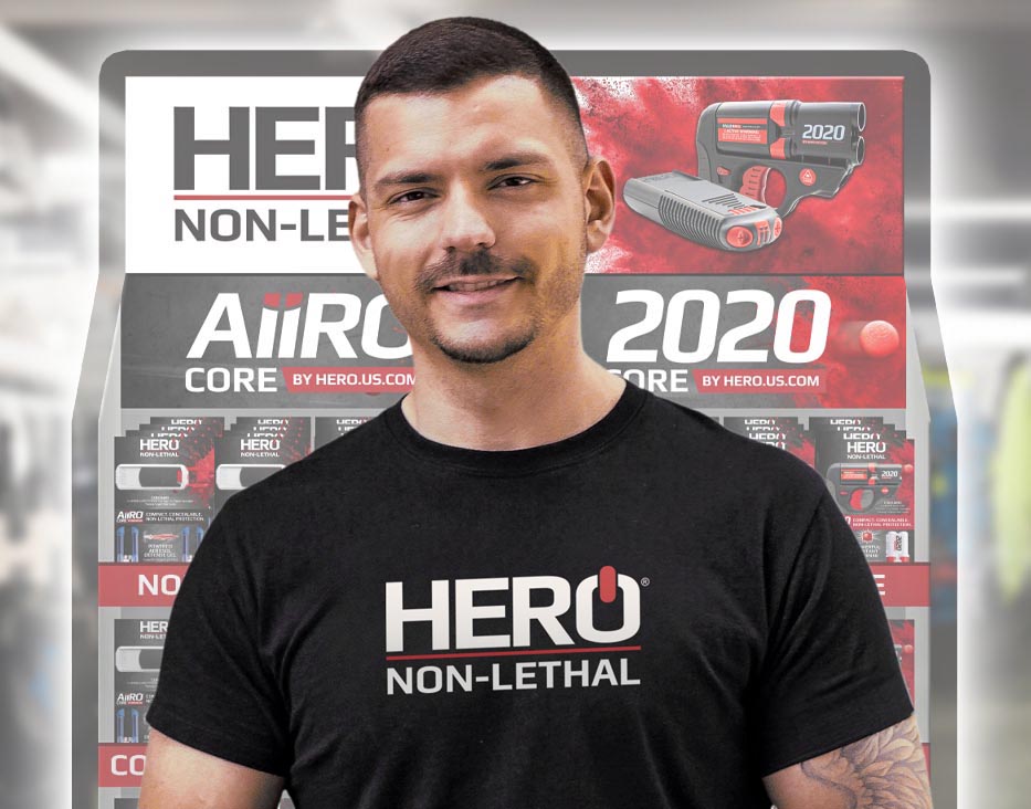 A man in a HERO Non-Lethal shirt stands in front of a retail display of AIIRO and HERO 2020 products